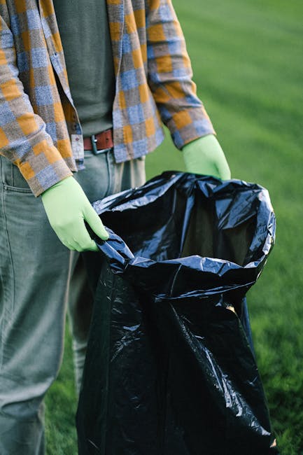 A person wearing a white glove is holding a large white polypropylene sack filled with garden debris, including small branches, leaves, and twigs, some of which are intertwined with green ivy. The sac