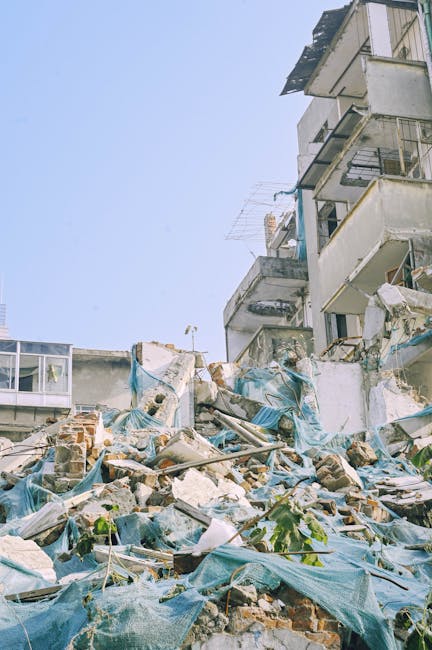 A scene of building debris and rubble outdoors at the base of a partially damaged multi-storey residential building. The foreground features broken bricks, concrete fragments, and scattered constructi